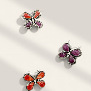 Three butterfly-shaped jewelry pendants with silver-toned filigree borders and cabochon stones, arranged on a light beige background. Two pendants feature orange-red stones, and one features purple stones.