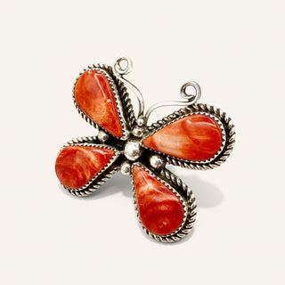 A butterfly-shaped silver pendant featuring four carved orange-red stone wings, set with small silver beads and a twisted rope border, on a light cream background.